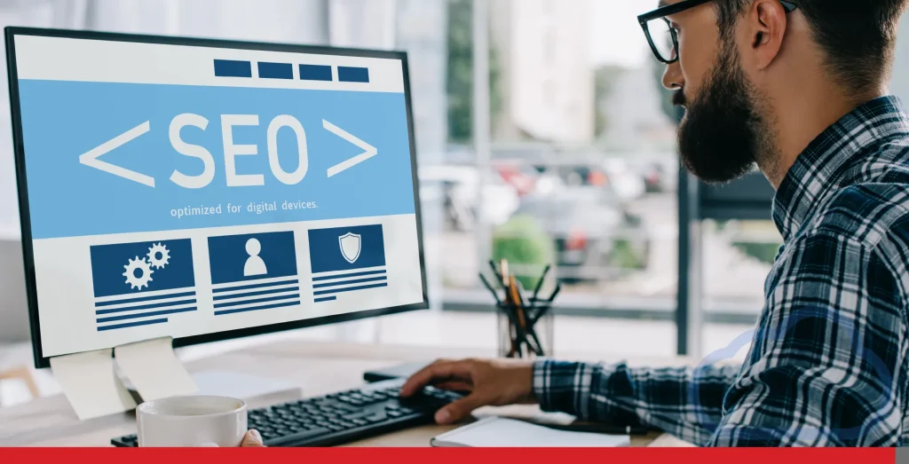 Young successful developer sitting at workplace with SEO sign on computer screen.